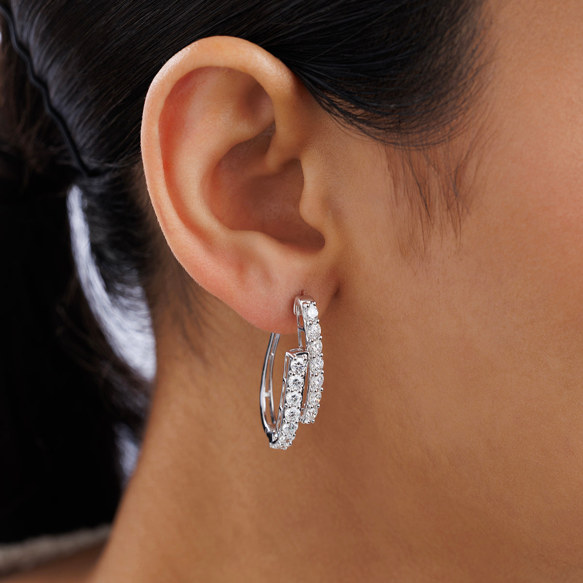 Close-up of a woman's ear wearing SOLACE Arc Huggie EARRINGS by Keemti Jewels, featuring double curved hoops encrusted with sparkling stones in a silver-tone setting. These earrings are a stunning everyday essential with a softly blurred background.