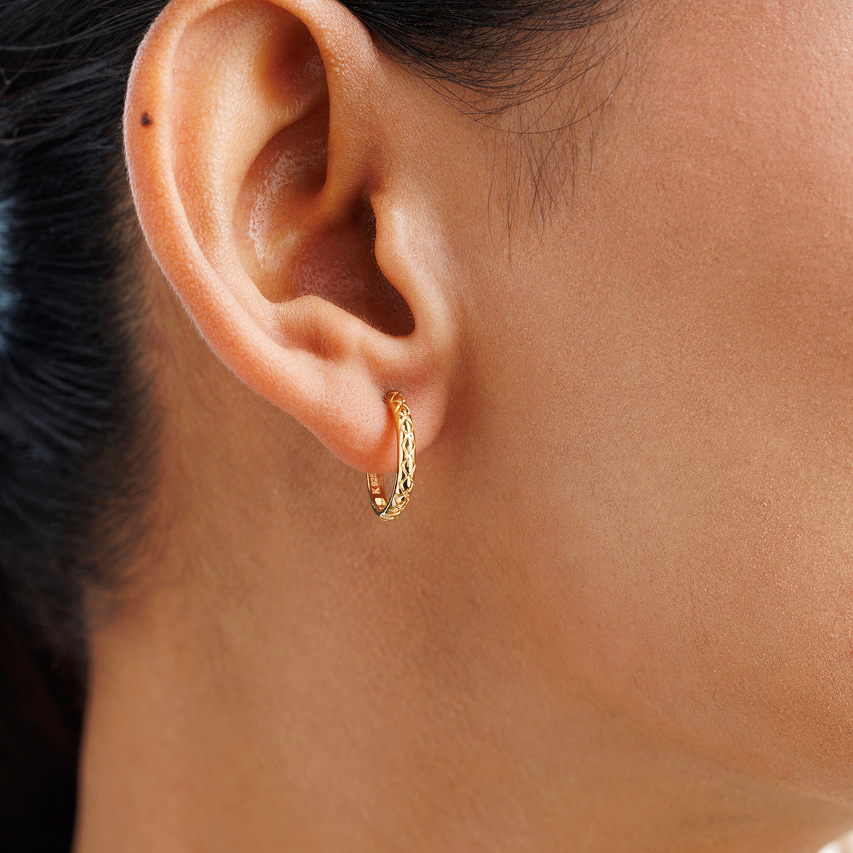 Close-up of a medium-skinned woman wearing Keemti Jewels' AURA Light Hoop EARRINGS, small gold hoops with a subtle braided design. The softly blurred background highlights this elegant jewelry piece.