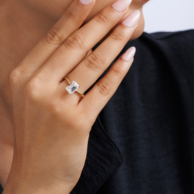 A hand with light pink nails wears the GRACE Emerald Solitaire Ring by Keemti Jewels, touching the chin.