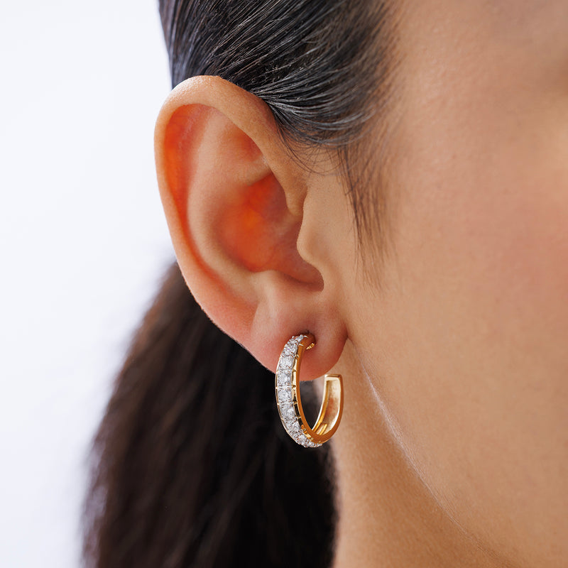 A close-up of a woman's ear showcases Keemti Jewels' CLASSIC Curve Hoop EARRINGS, featuring yellow plating and sparkling round diamonds. The light, blurred background highlights the elegance of the huggie hoops.