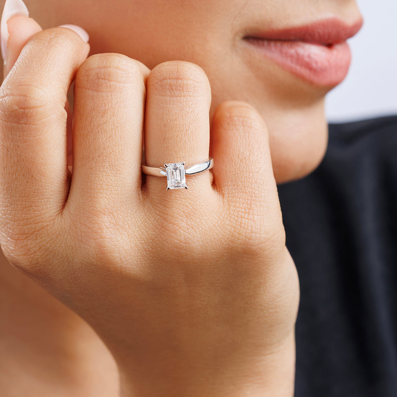A hand with the MINIMAL Emerald Solitaire RING by Keemti Jewels, featuring a silver band, near their face.