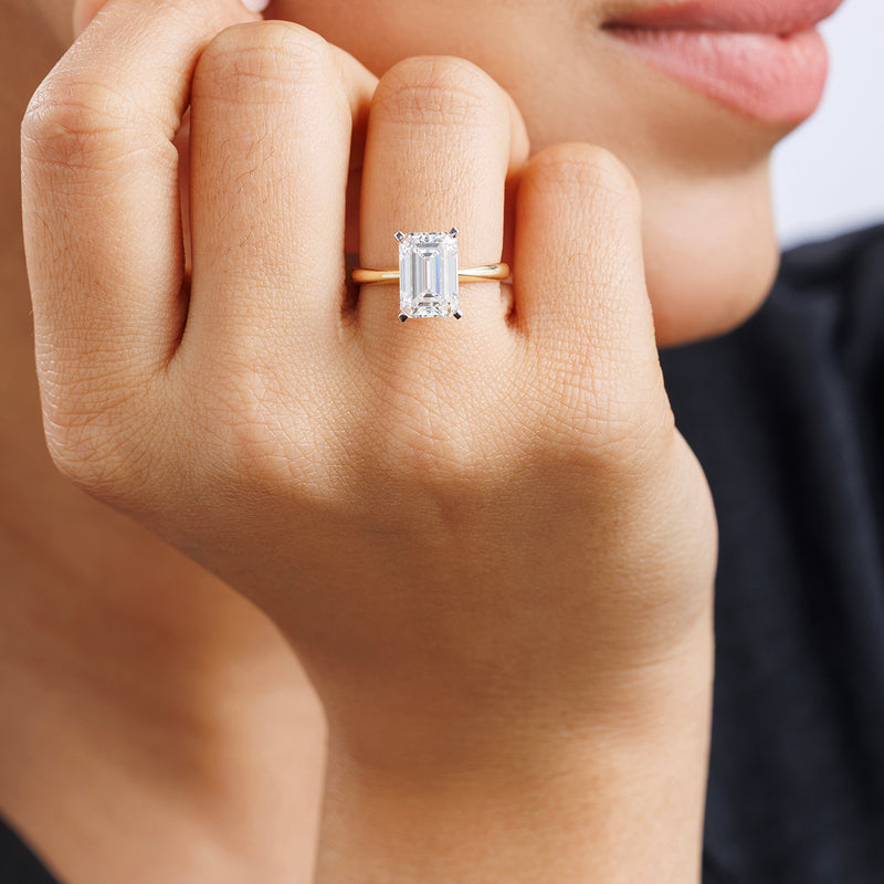 A woman's hand wears the Keemti Jewels CLASSIC Emerald Solitaire RING on a gold band, held near her smiling lips.