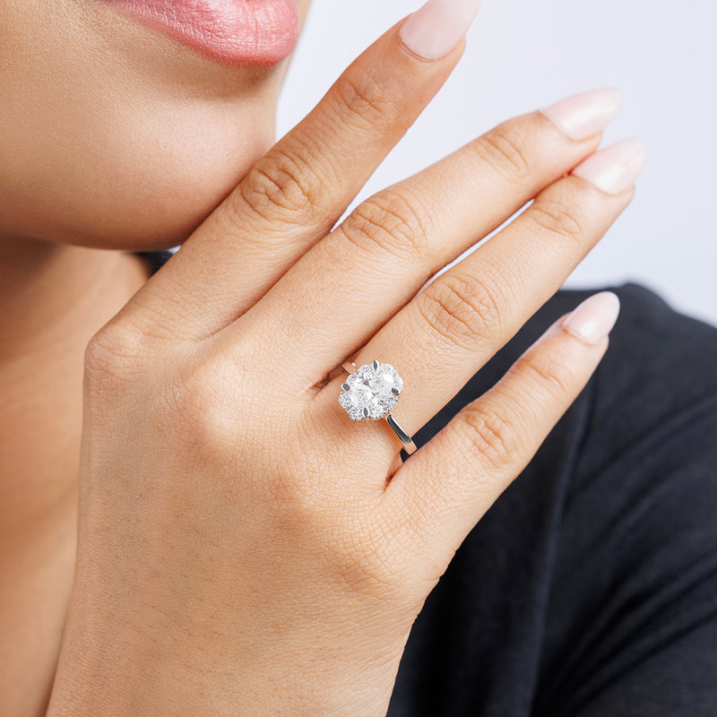A woman with light pink nails gently touches her face, wearing the Keemti Jewels CLASSIC Oval Mount Halo Ring.