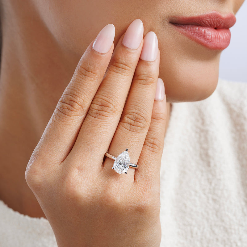 A woman with manicured nails highlights the Keemti Jewels TIMELESS Pear Solitaire RING as she touches her face.