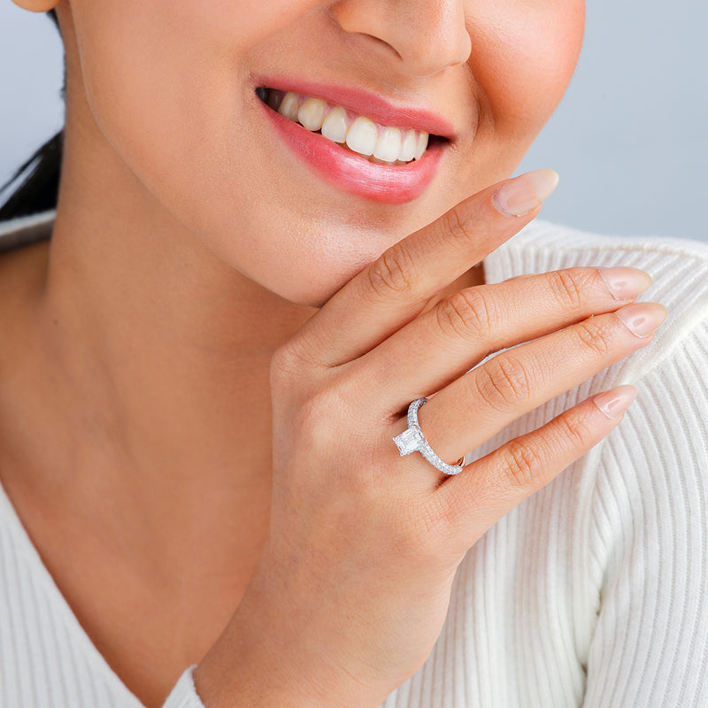 A smiling woman in a white top displays the Keemti Jewels Timeless Emerald Solitaire Ring near her face.